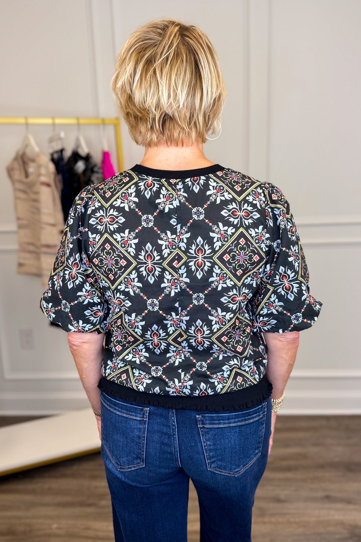 Back view of woman wearing Swagger Boutique’s Black Embroidered Print Top with jeans with clothing hanging in the background.