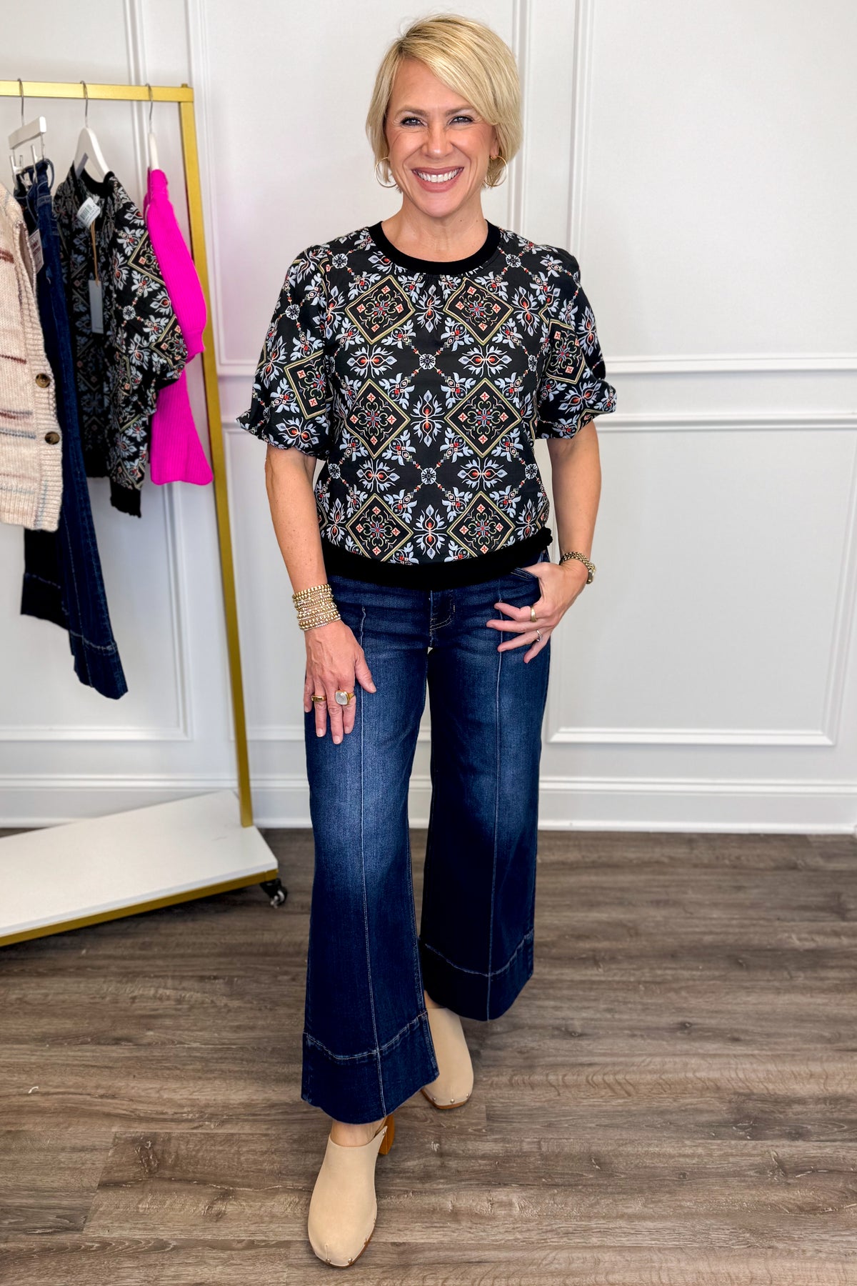 Front view of woman wearing Swagger Boutique’s Black Embroidered Print Top with jeans with clothing hanging in the background.