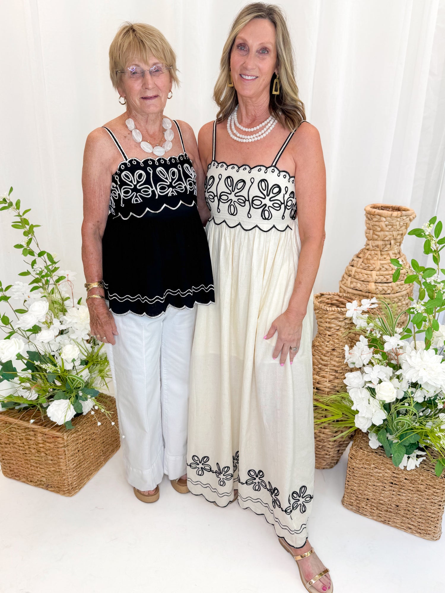Front view of woman wearing Swagger Boutique’s Black &amp; Ivory Embroidered Tank with jeans with daughter wearing Cream &amp; Black Embroidered Maxi Dress against white studio background.