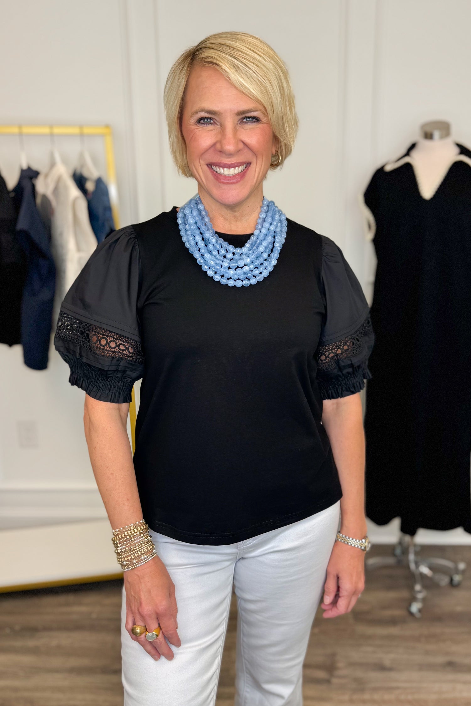 Front view of woman wearing Swagger Boutique’s Black Lace Sleeve Top with jeans with clothing hanging in the background.