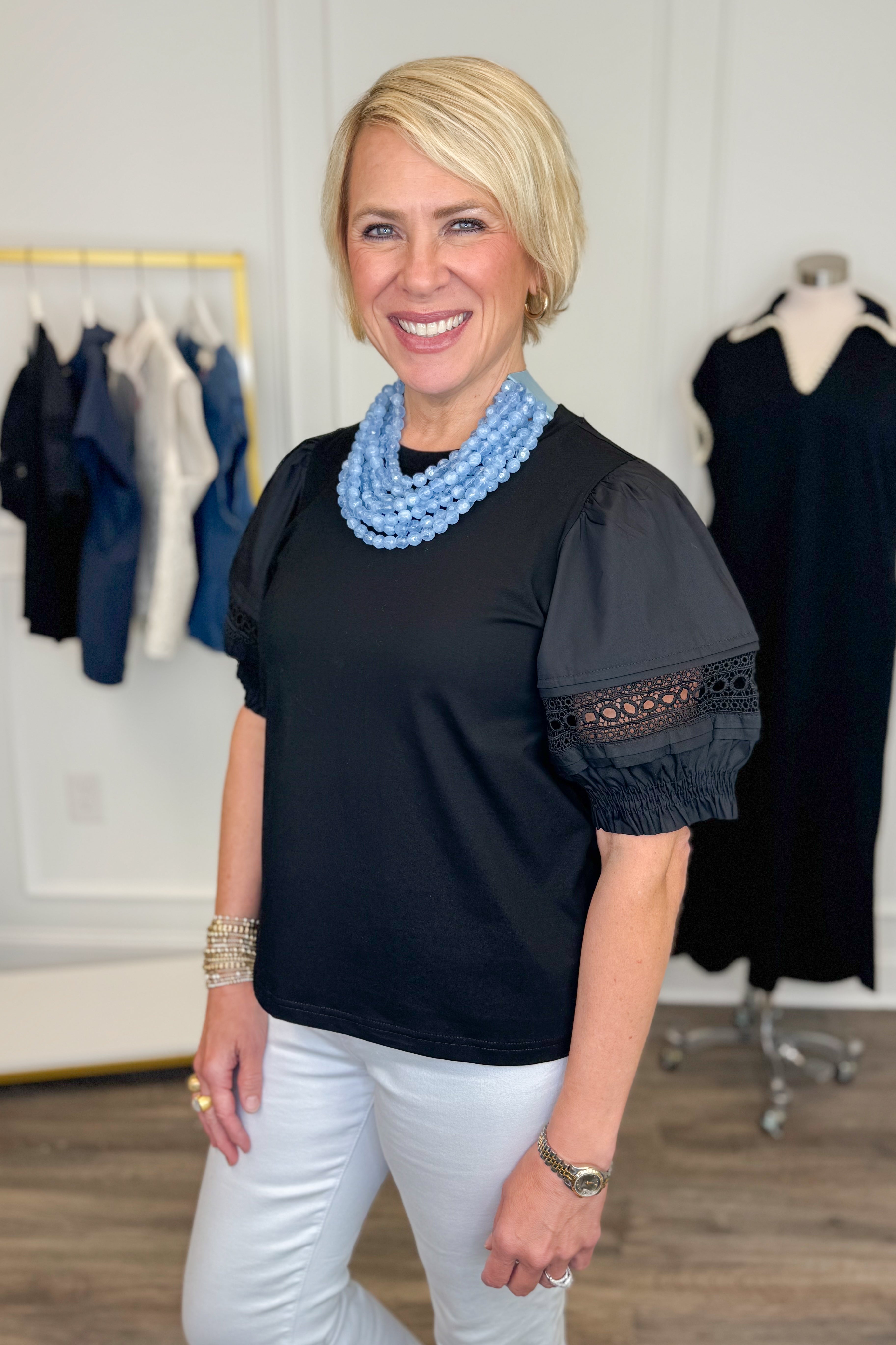 Side view of woman wearing Swagger Boutique’s Black Lace Sleeve Top with jeans with clothing hanging in the background.