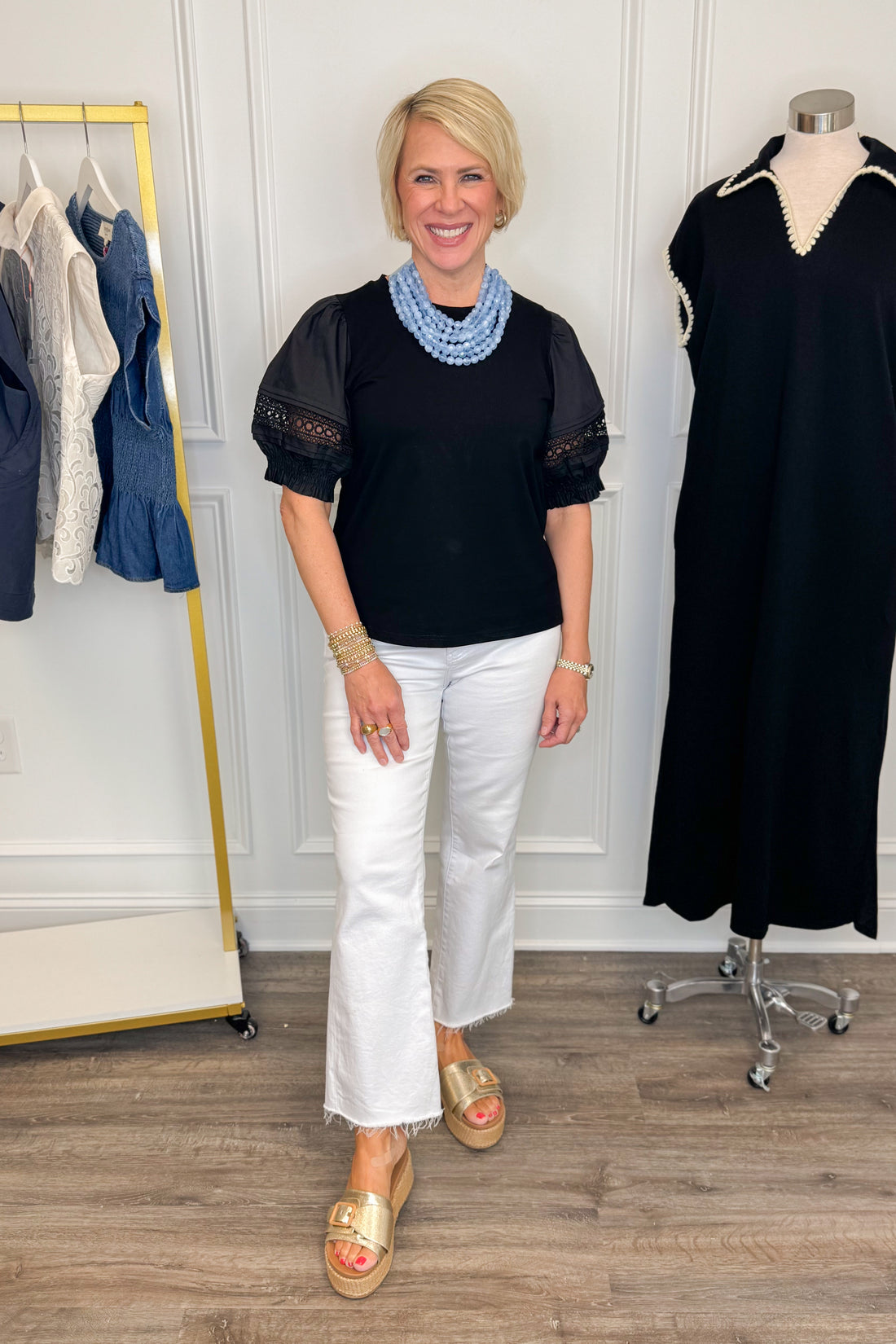 Front view of woman wearing Swagger Boutique’s Black Lace Sleeve Top with jeans with clothing hanging in the background.