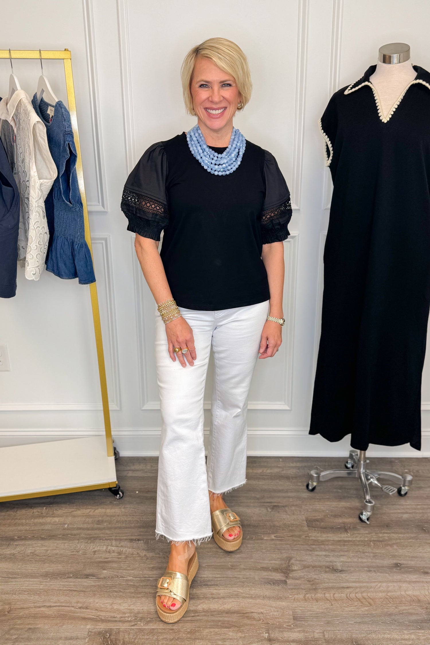 Front view of woman wearing Swagger Boutique’s Black Lace Sleeve Top with jeans with clothing hanging in the background.