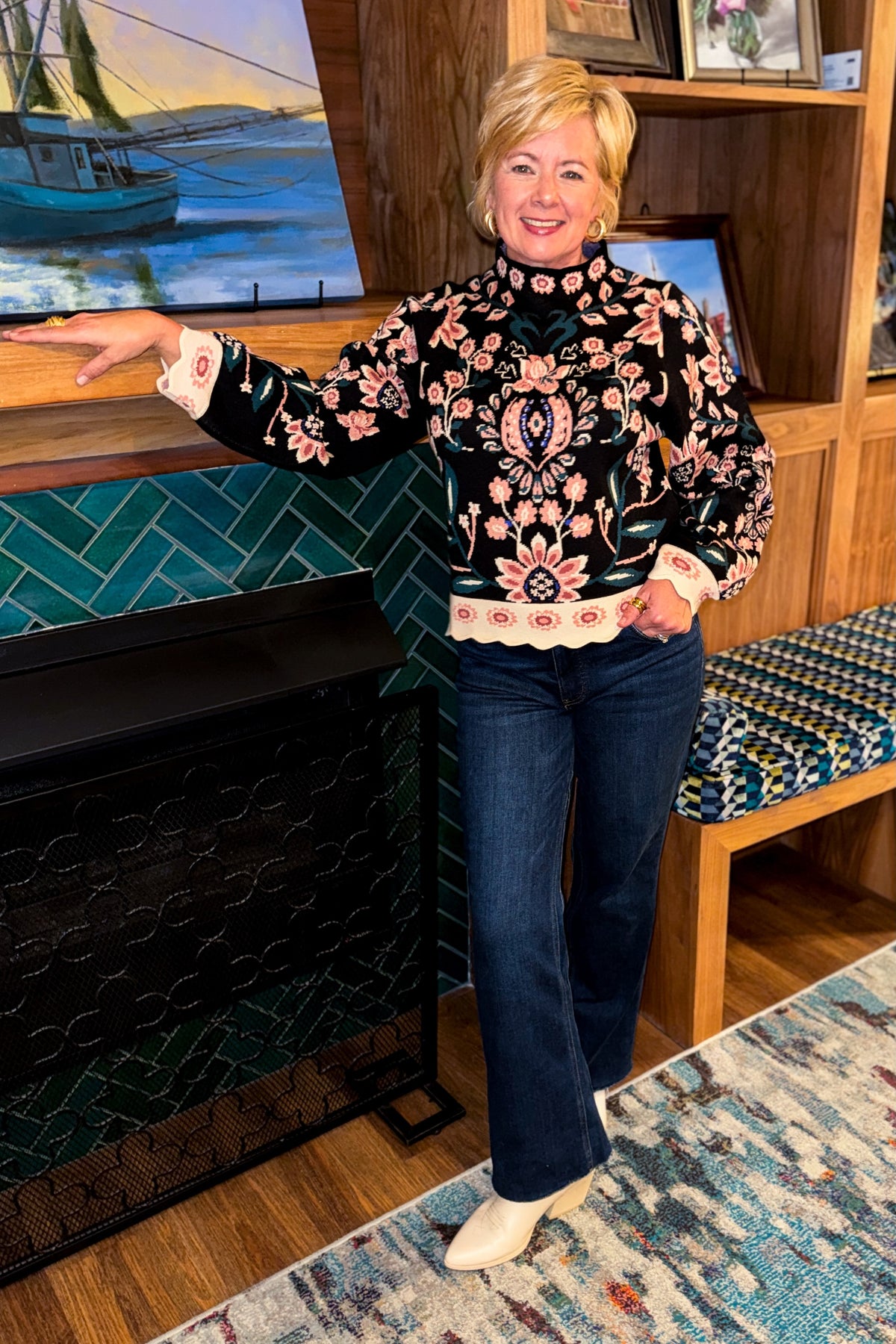 Front view of woman wearing Swagger Boutique’s Black &amp; Multi Floral Print Sweater by THML with jeans standing by a fireplace. 