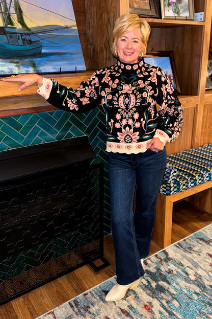 Front view of woman wearing Swagger Boutique’s Black &amp; Multi Floral Print Sweater by THML with jeans standing by a fireplace. 