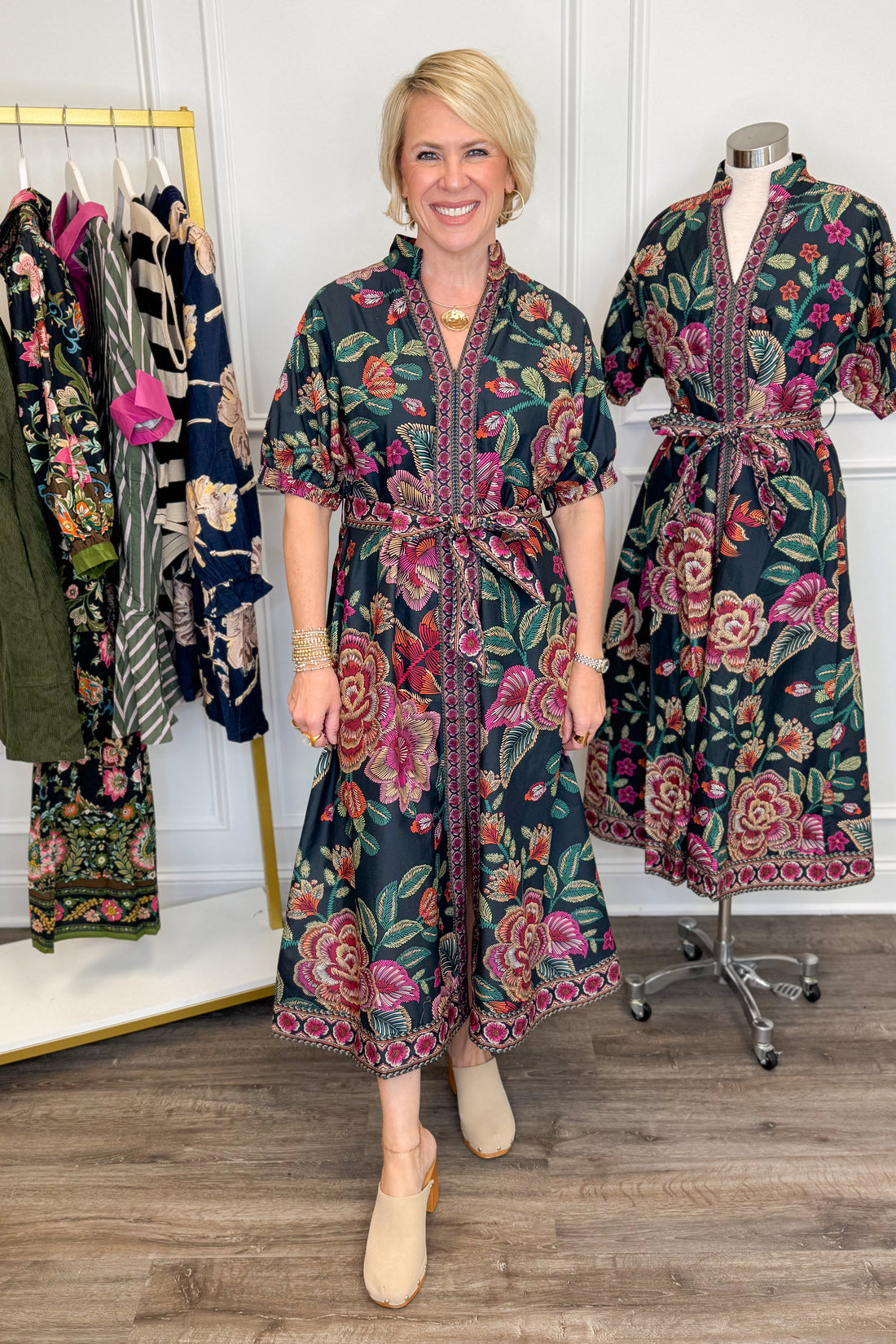Front view of woman wearing Swagger Boutique’s Black &amp; Multi Printed 3/4 Sleeve Midi Dress by Entro with clogs with clothing hanging in the background.
