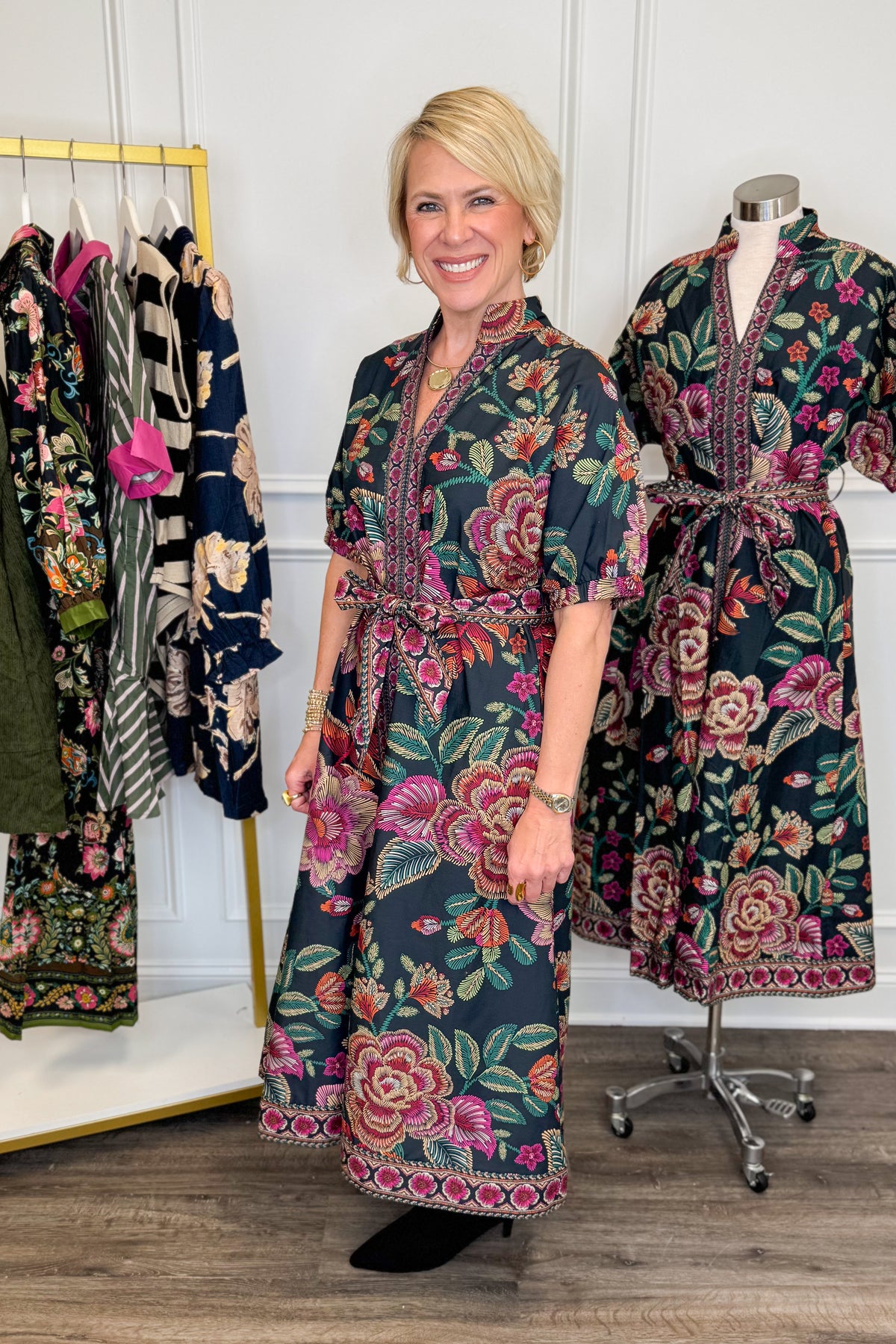 Side view of woman wearing Swagger Boutique’s Black &amp; Multi Printed 3/4 Sleeve Midi Dress by Entro with boots with clothing hanging in the background.