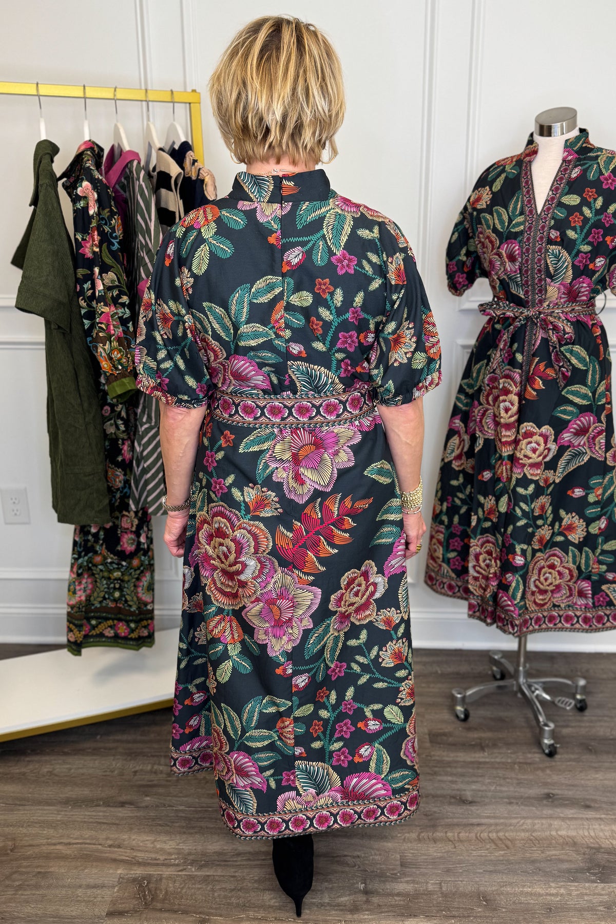 Back view of woman wearing Swagger Boutique’s Black &amp; Multi Printed 3/4 Sleeve Midi Dress by Entro with boots with clothing hanging in the background.