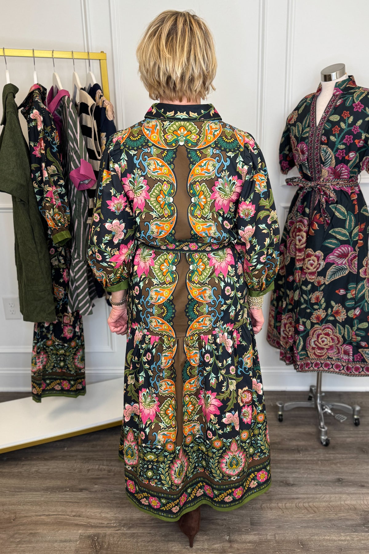 Back view of woman wearing Swagger Boutique’s black multi printed midi dress by Entro with boots with clothing hanging in the background.