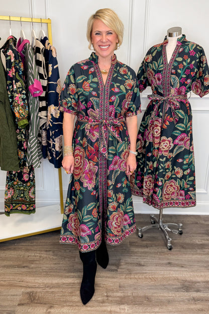 Front view of woman wearing Swagger Boutique’s Black &amp; Multi Printed 3/4 Sleeve Midi Dress by Entro with boots with clothing hanging in the background.
