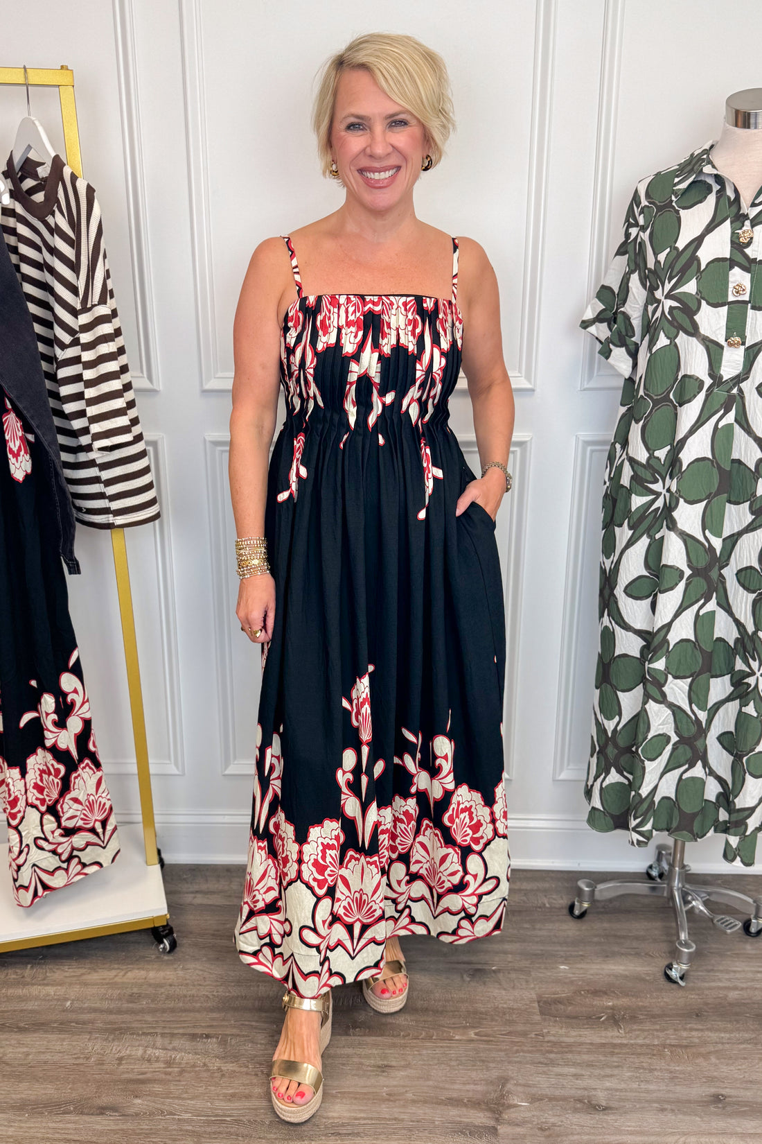 Front view of woman wearing Swagger Boutique’s Black &amp; Red Floral Midi Dress by Entro with sandals with clothing hanging in the background. 