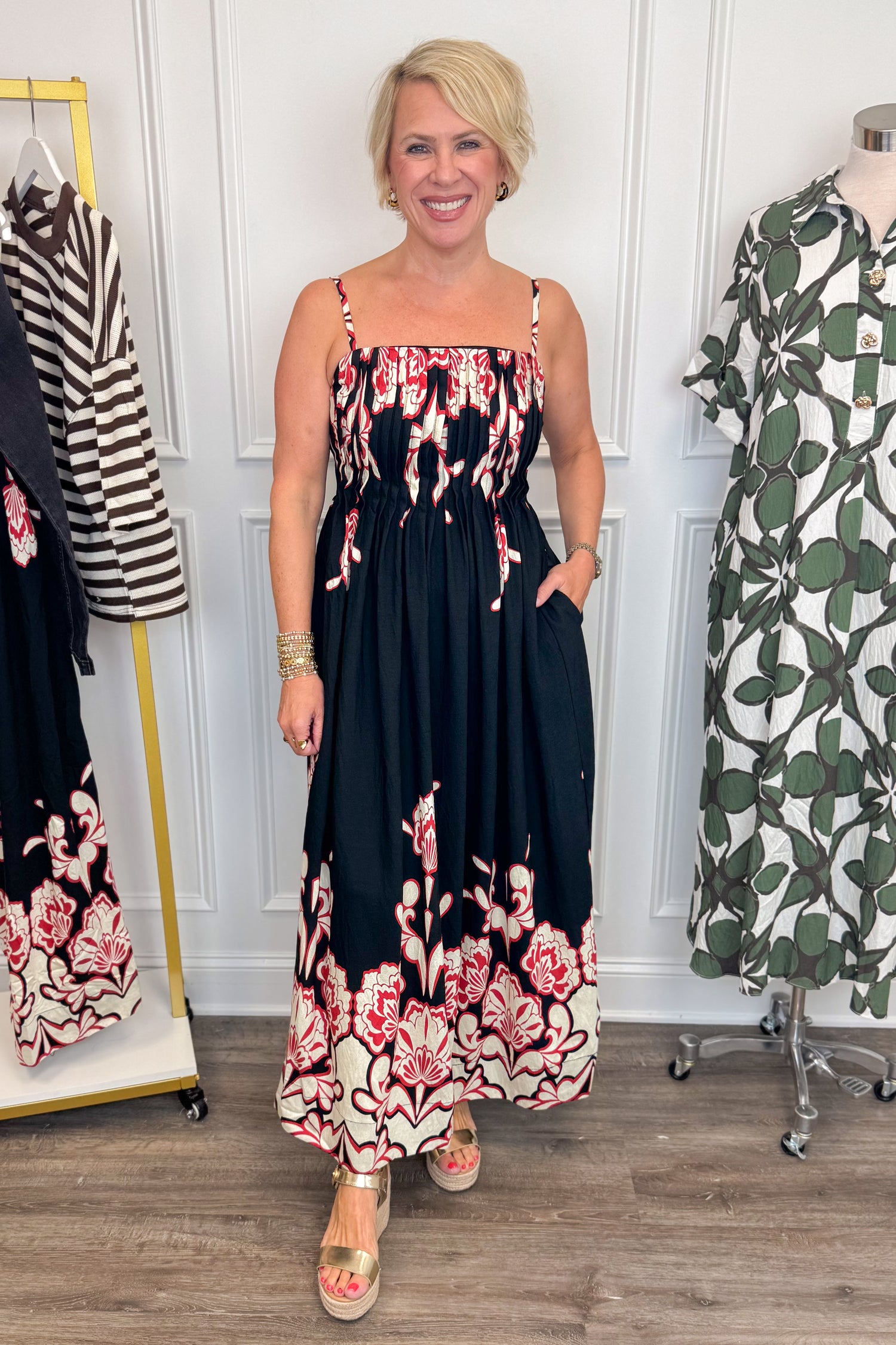 Front view of woman wearing Swagger Boutique’s Black &amp; Red Floral Midi Dress by Entro with sandals with clothing hanging in the background. 