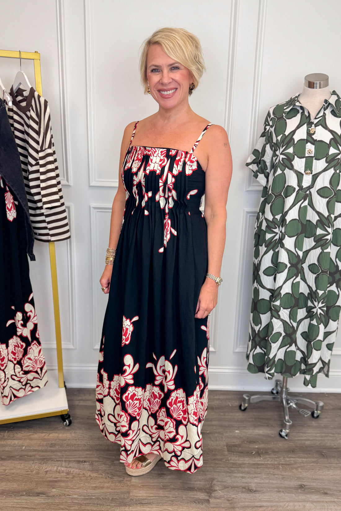 Side view of woman wearing Swagger Boutique’s Black &amp; Red Floral Midi Dress by Entro with sandals with clothing hanging in the background. 