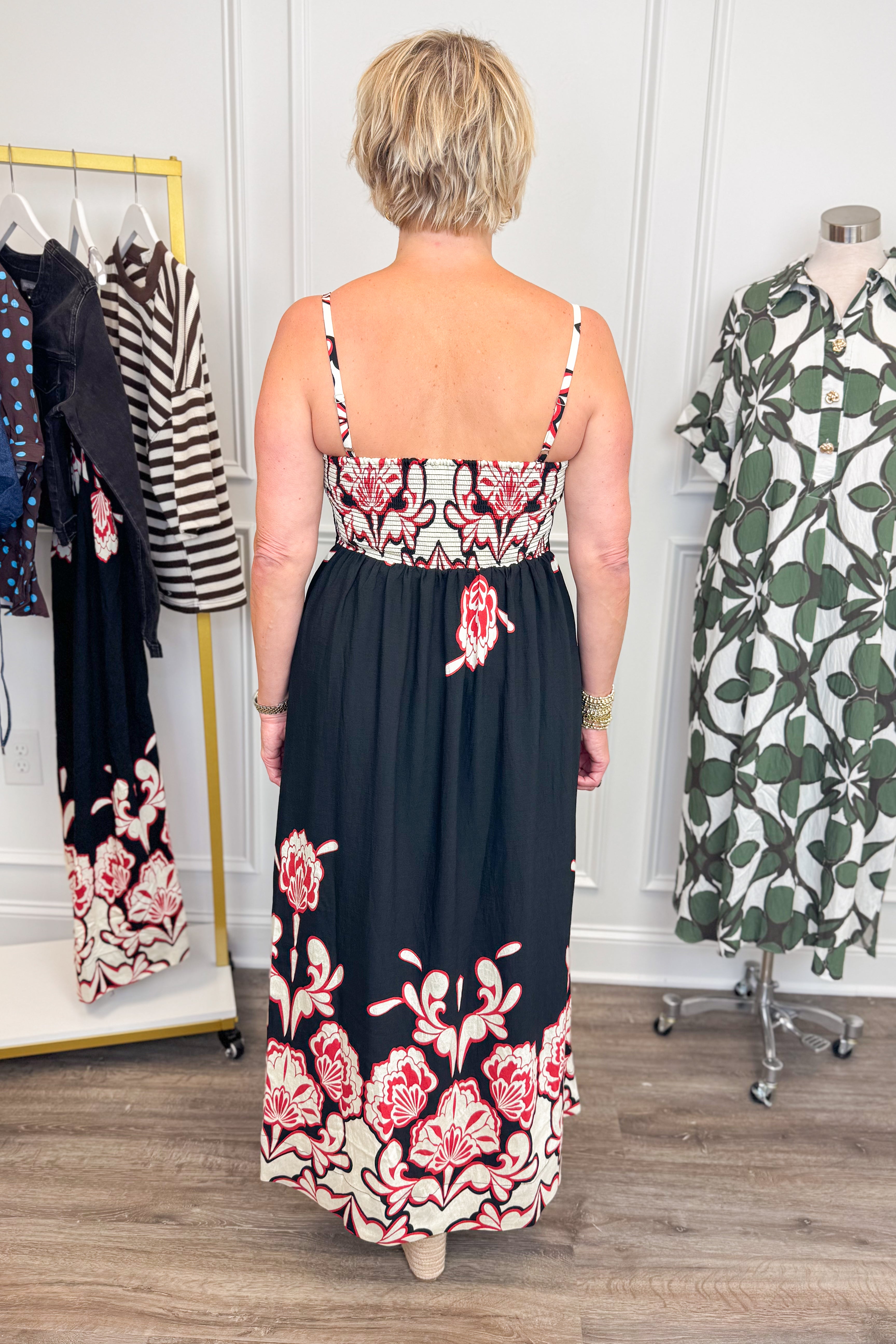 Back view of woman wearing Swagger Boutique’s Black &amp; Red Floral Midi Dress by Entro with sandals with clothing hanging in the background. 