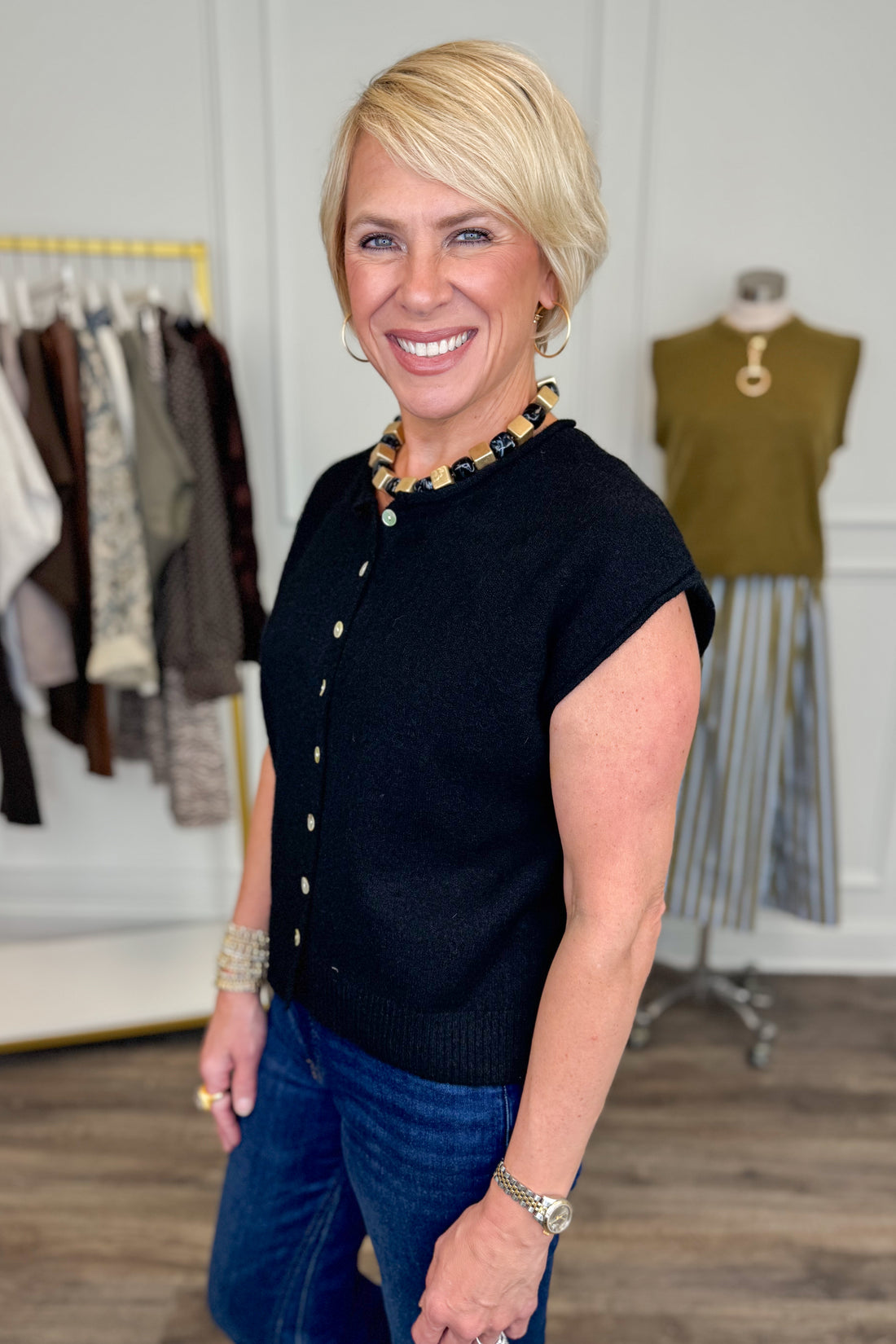 Side view of woman wearing Swagger Boutique’s Black Sleeveless Button Up Sweater with jeans with clothing hanging in the background.