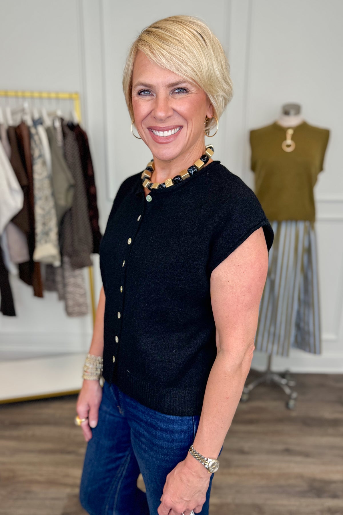 Side view of woman wearing Swagger Boutique’s Black Sleeveless Button Up Sweater with jeans with clothing hanging in the background.