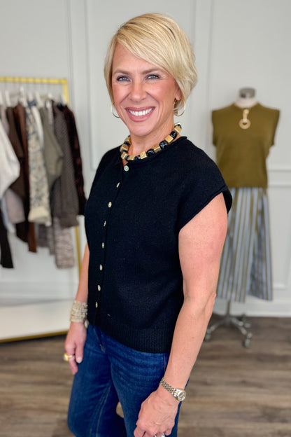 Side view of woman wearing Swagger Boutique’s Black Sleeveless Button Up Sweater with jeans with clothing hanging in the background.