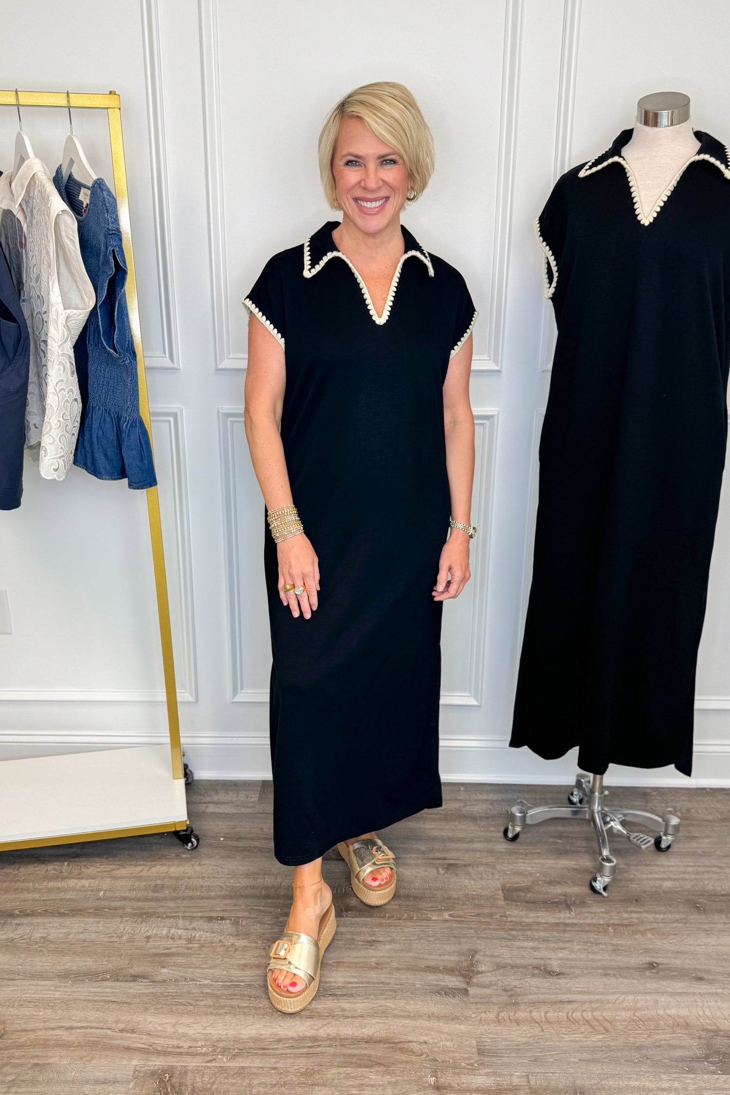 Front view of woman wearing Swagger Boutique’s Black Whipstitch Midi Dress by Entro with sandals with clothing hanging in the background.