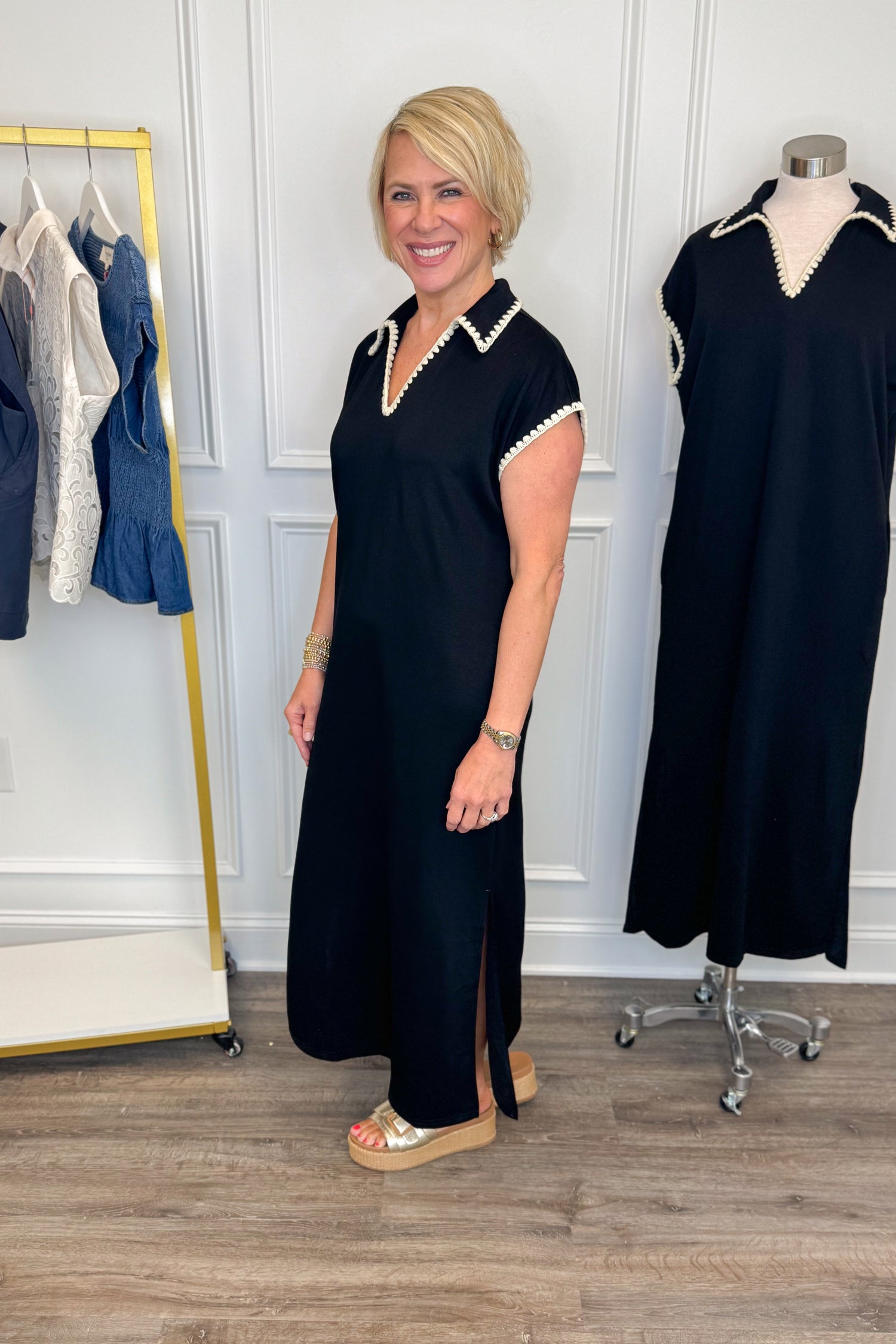 Side view of woman wearing Swagger Boutique’s Black Whipstitch Midi Dress by Entro with sandals with clothing hanging in the background.