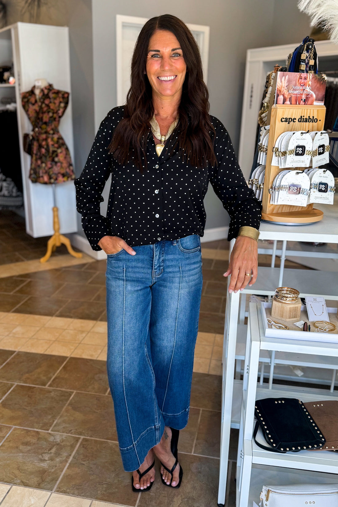 Front view of woman wearing Swagger Boutique’s Black &amp; White Polka Dot Satin Button Up Top with jeans standing at the store.