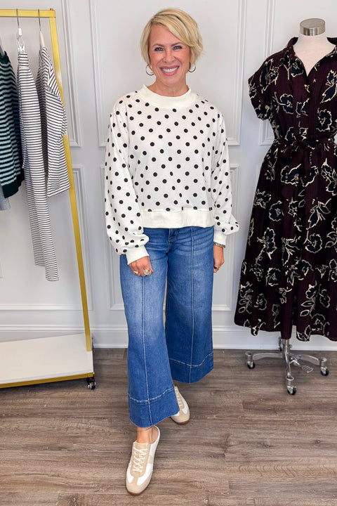 Front view of woman wearing Swagger Boutique’s Black &amp; White Polka Dot Sweatshirt with jeans with clothing hanging in the background. 
