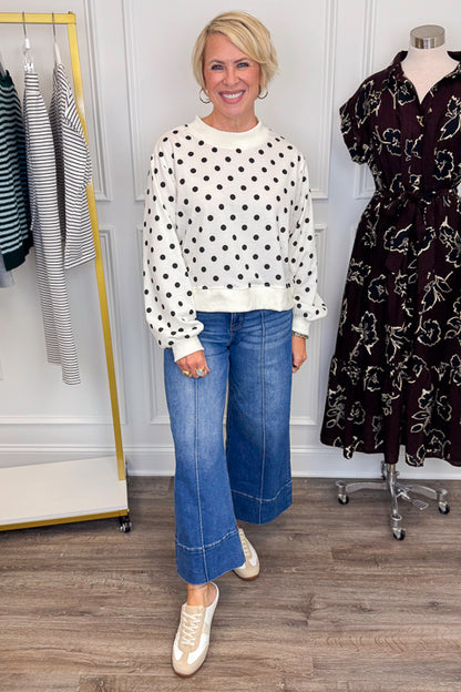 Front view of woman wearing Swagger Boutique’s Black &amp; White Polka Dot Sweatshirt with jeans with clothing hanging in the background. 