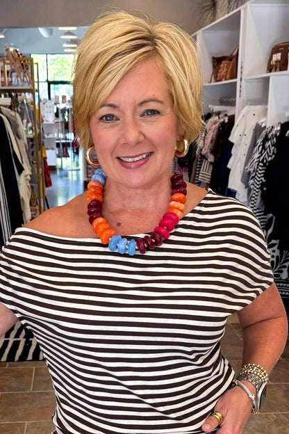 A smiling blonde woman wearing a colorful chunky beaded necklace and a black and white striped top, standing inside a clothing store.