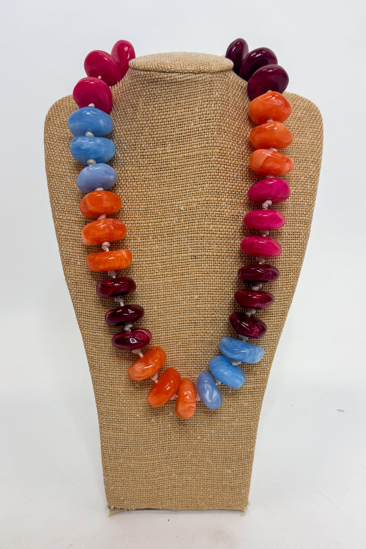 A chunky multi-colored beaded necklace displayed on a burlap-covered jewelry bust. The necklace features irregular polished beads in bright magenta pink, deep burgundy, orange, and sky blue, separated by small silver spacer beads.