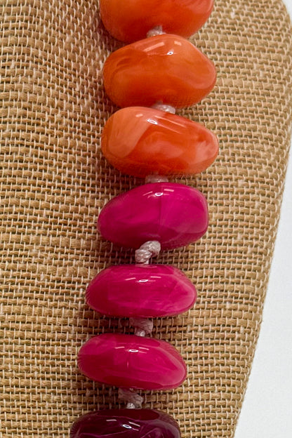 A close-up of a beaded strand with large polished oval beads in orange, magenta pink, and deep burgundy, displayed on a tan burlap surface.