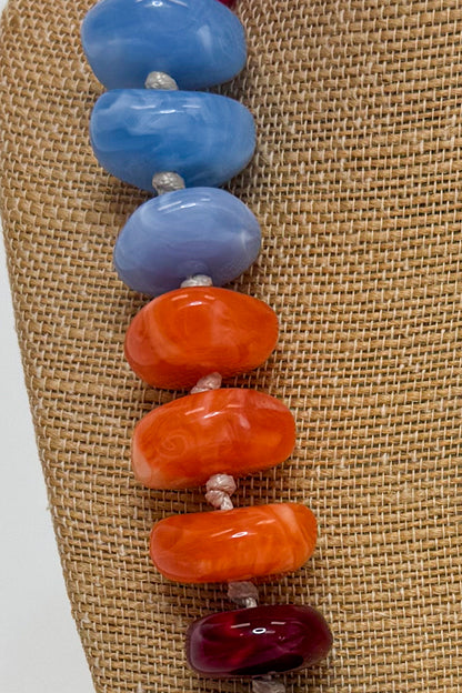 A close-up of a beaded strand with large oval glass beads in light blue, lavender, bright orange, and deep burgundy, threaded on a light cord against a tan woven fabric background.