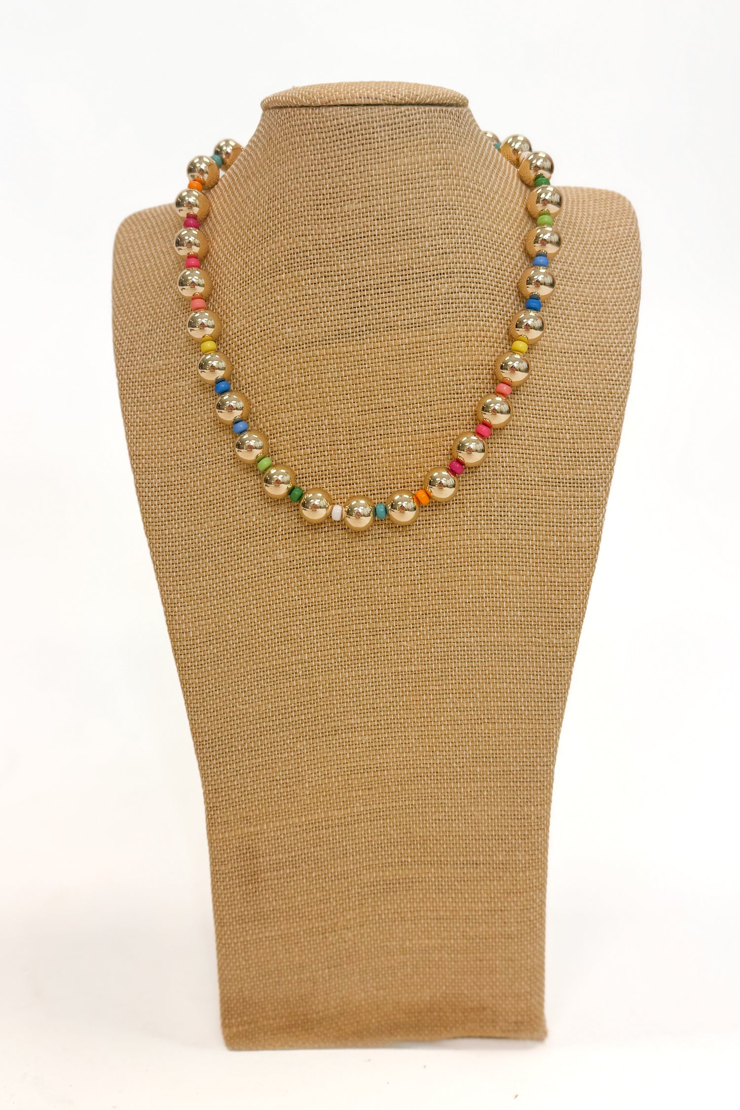 a gold and colorful beaded necklace on a burlap necklace stand with a white background