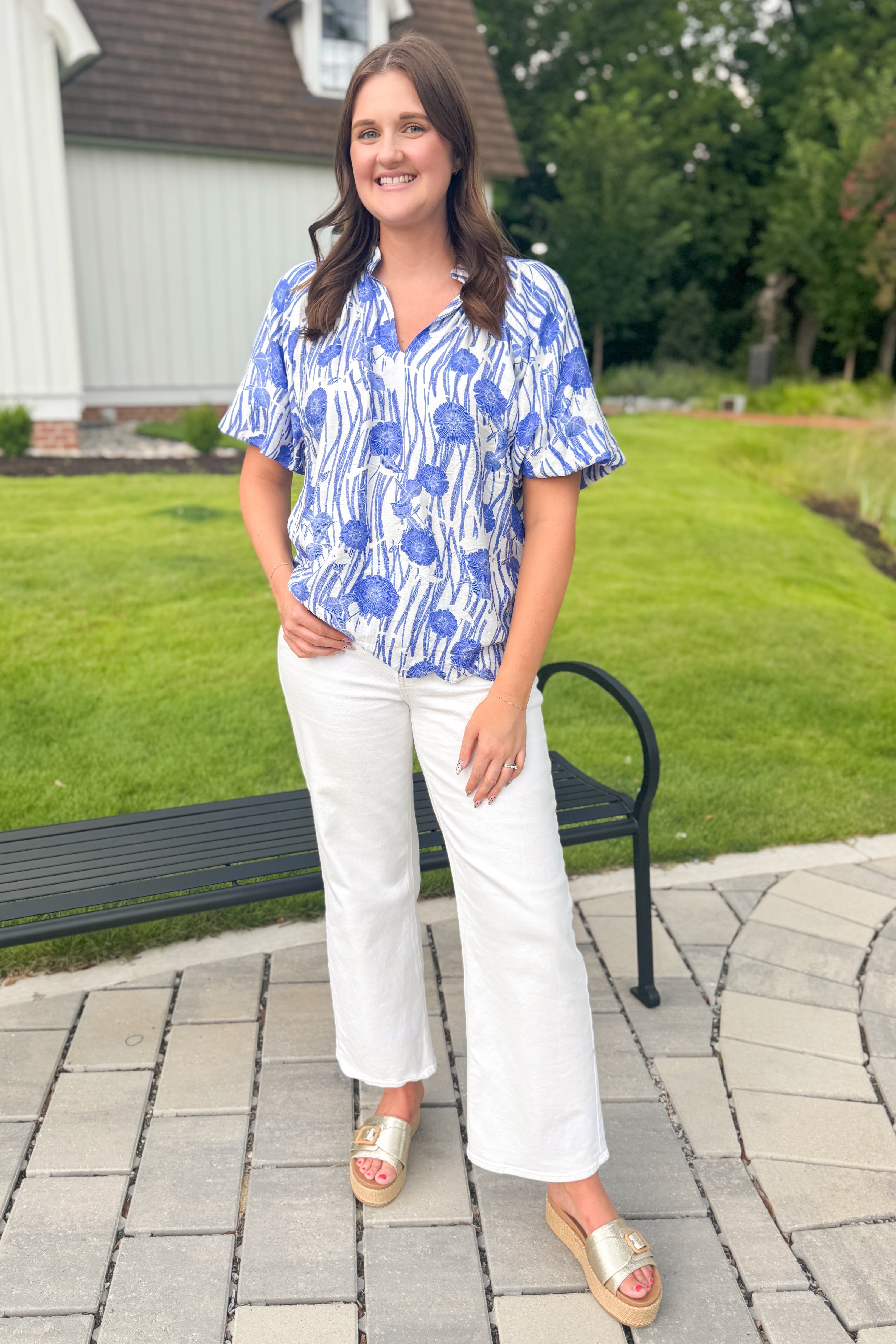 Front view of woman wearing Swagger Boutique’s Blue Floral Puff Sleeve Top with jeans standing outside by a bench.