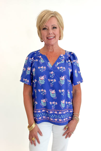 Front view of woman wearing Swagger Boutique’s Blue Petal Print Lori Top by Mary Square with white jeans against white studio background.