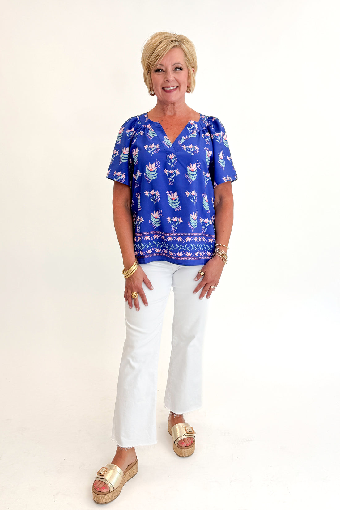 Front view of woman wearing Swagger Boutique’s Blue Petal Print Lori Top by Mary Square with white jeans against white studio background.