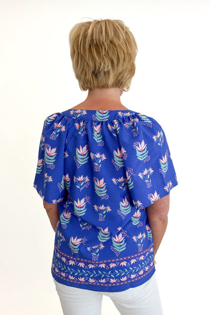 Back view of woman wearing Swagger Boutique’s Blue Petal Print Lori Top by Mary Square with white jeans against white studio background.