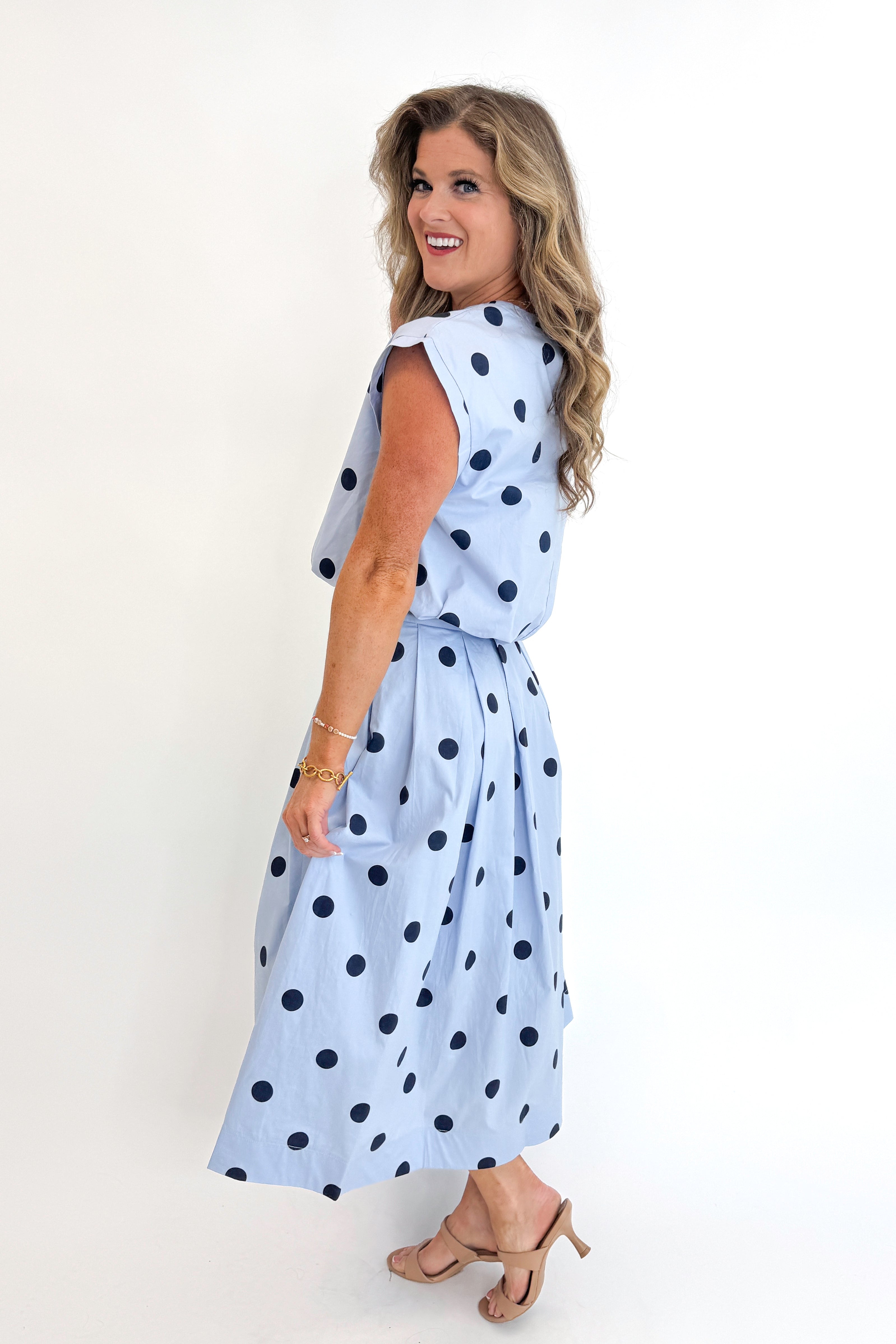 Side view of woman wearing Swagger Boutique’s Blue Polka Dot Bubble Hem Tank with matching Blue Polka Dot Pleated Midi Skirt against white studio background.