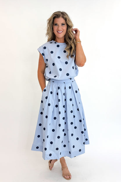 Side view of woman wearing Swagger Boutique’s Blue Polka Dot Bubble Hem Tank with matching Blue Polka Dot Pleated Midi Skirt against white studio background.