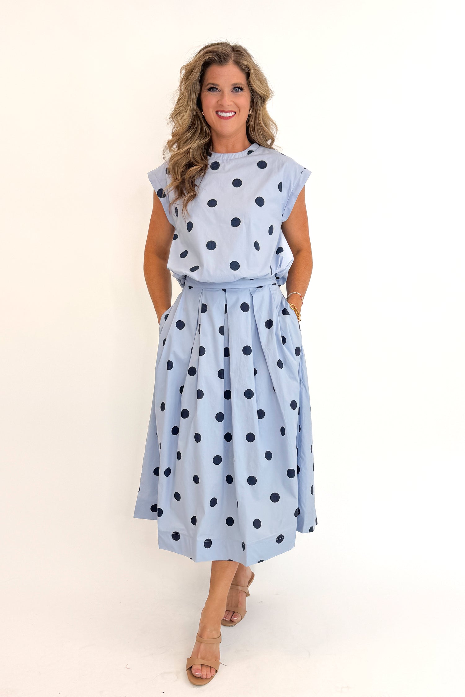 Front view of woman wearing Swagger Boutique’s Blue Polka Dot Bubble Hem Tank with matching Blue Polka Dot Pleated Midi Skirt against white studio background.