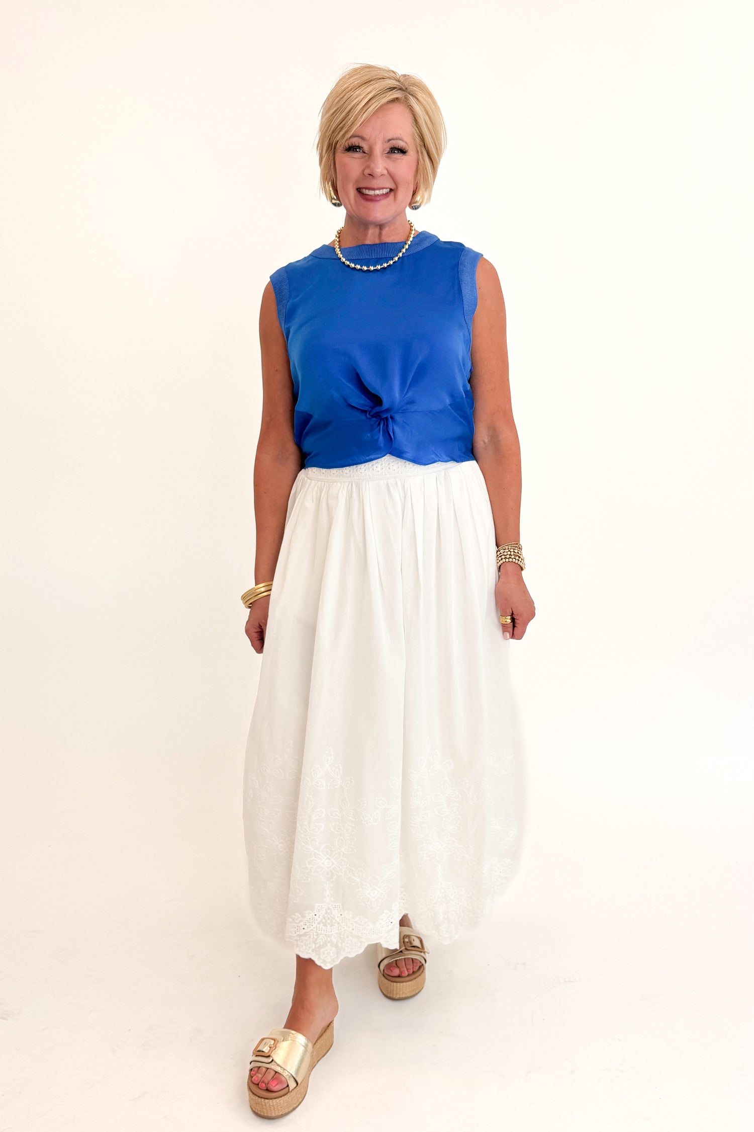 Front view of woman wearing Swagger Boutique’s Blue Sleeveless Tank With Twist by Skies Are Blue with White Embroidered Skirt against white studio background.