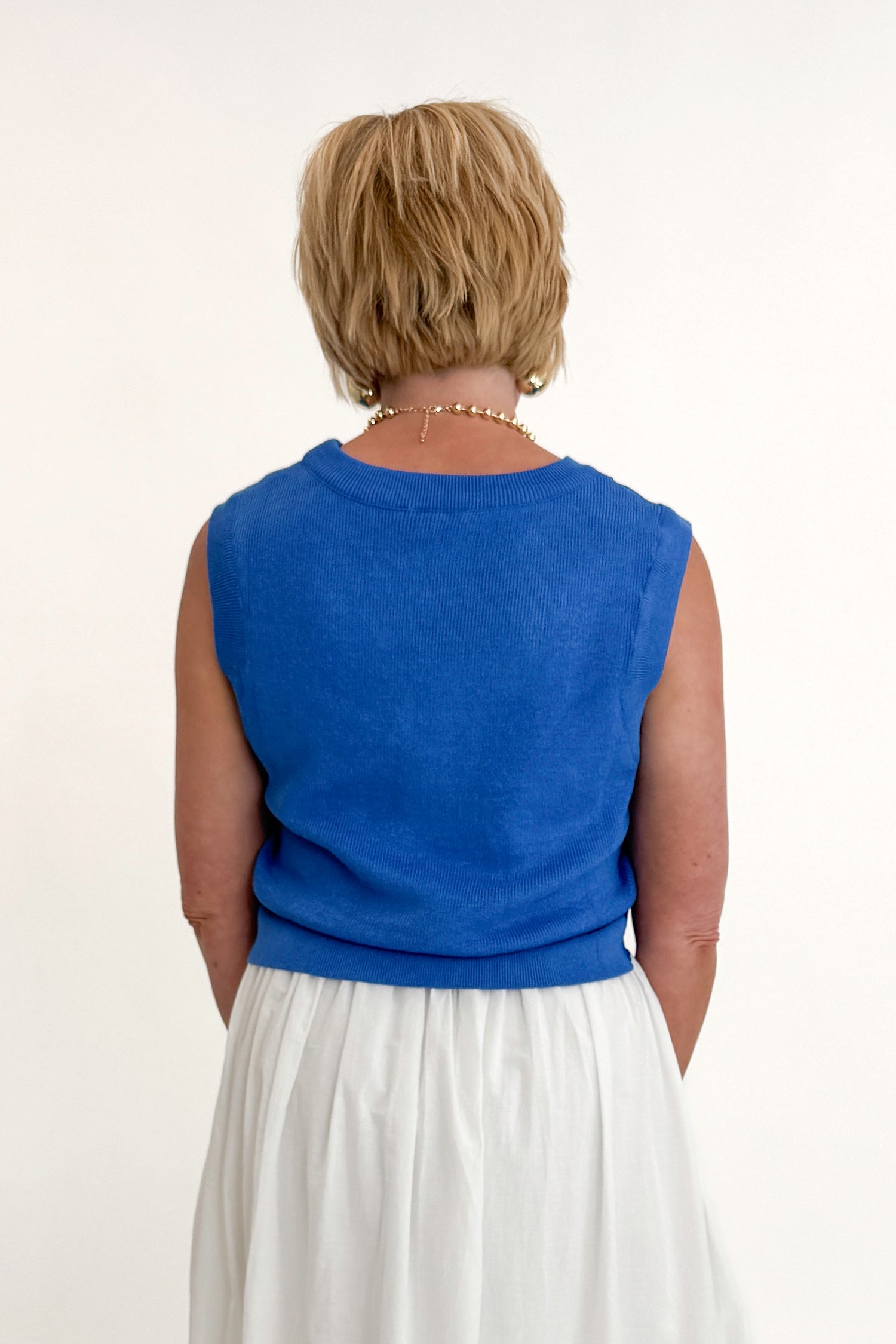 Back view of woman wearing Swagger Boutique’s Blue Sleeveless Tank With Twist by Skies Are Blue with white skirt against white studio background.