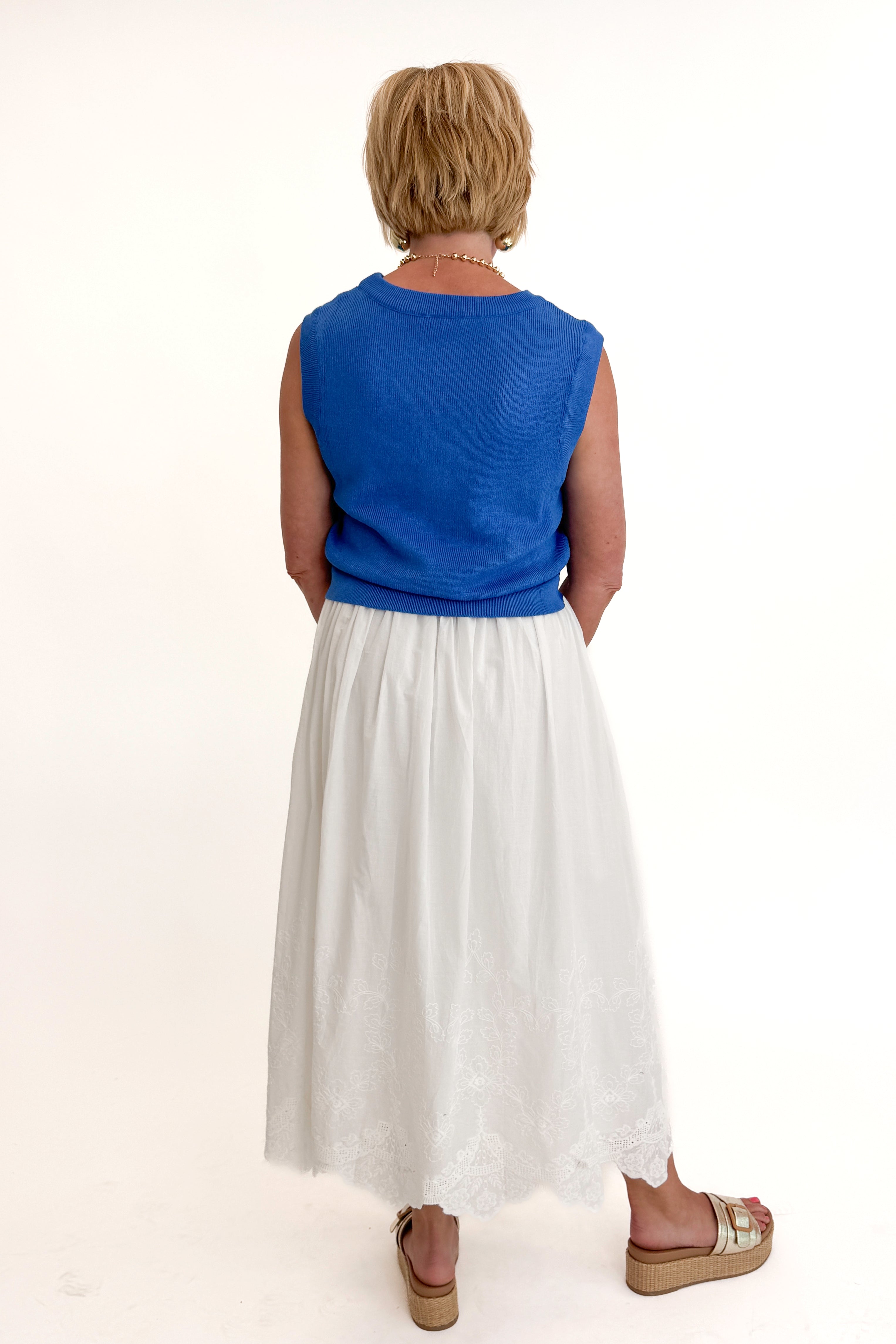 Back view of woman wearing Swagger Boutique’s Blue Sleeveless Tank With Twist by Skies Are Blue with White Embroidered Skirt against white studio background.
