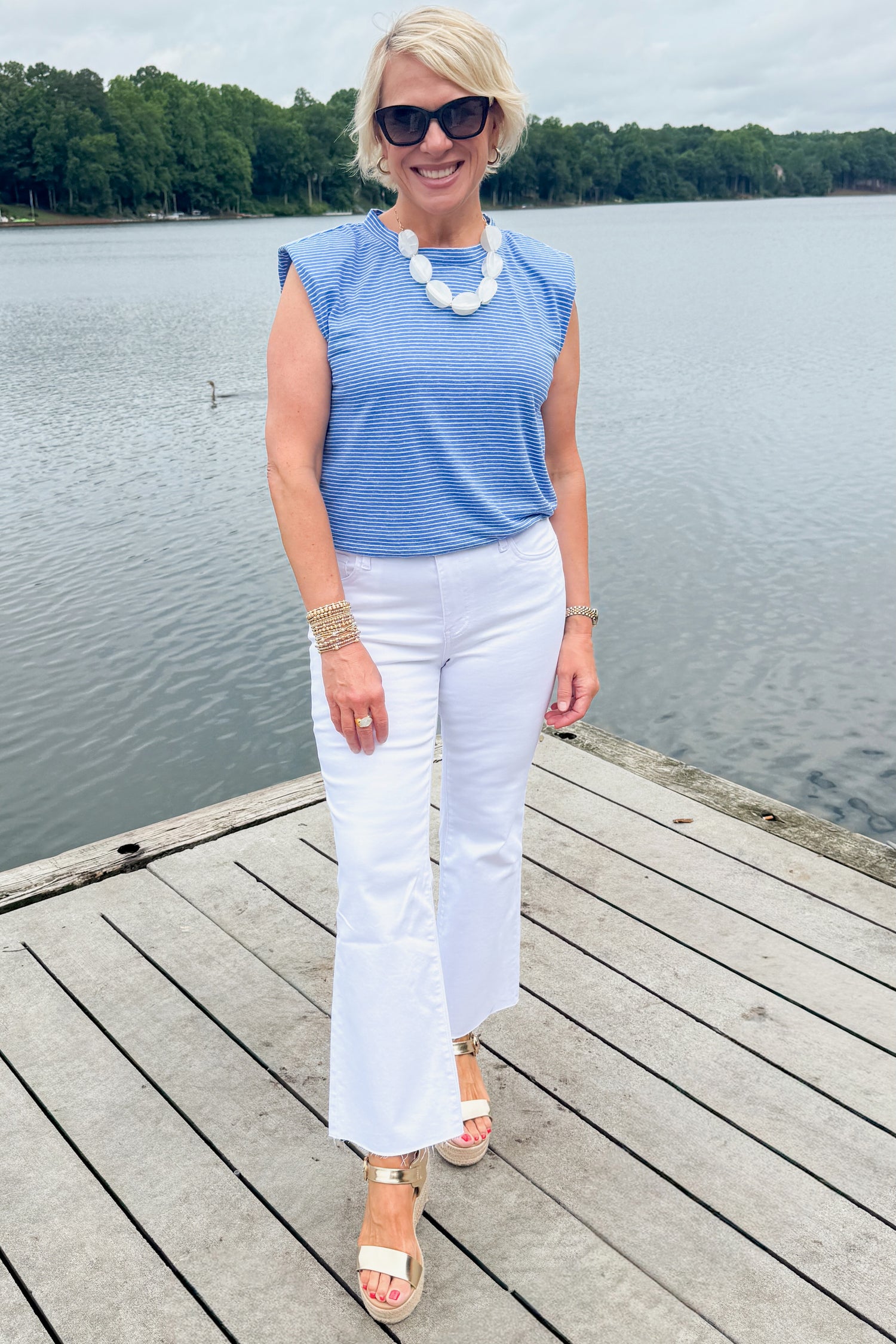 Front view of woman wearing Swagger Boutique’s Blue Striped Bubble Hem Sleeveless Top with jeans standing on a dock outside.