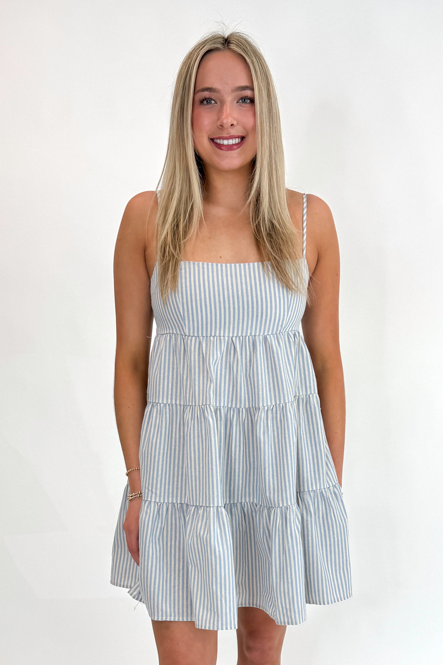 Front view of woman wearing Swagger Boutique’s Blue Striped Sleeveless Tiered Mini Dress with sandals against white studio background.