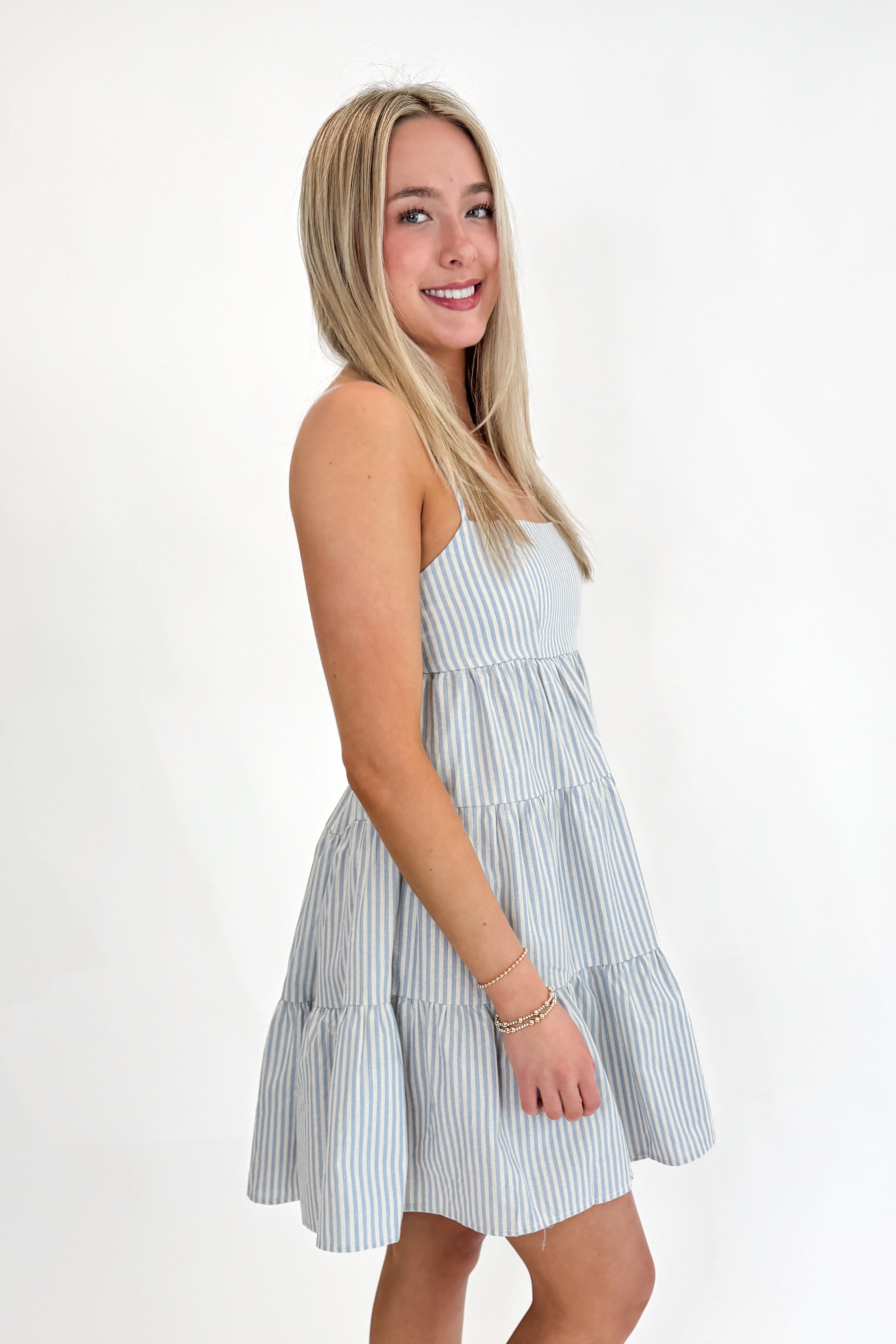 Side view of woman wearing Swagger Boutique’s Blue Striped Sleeveless Tiered Mini Dress with sandals against white studio background.