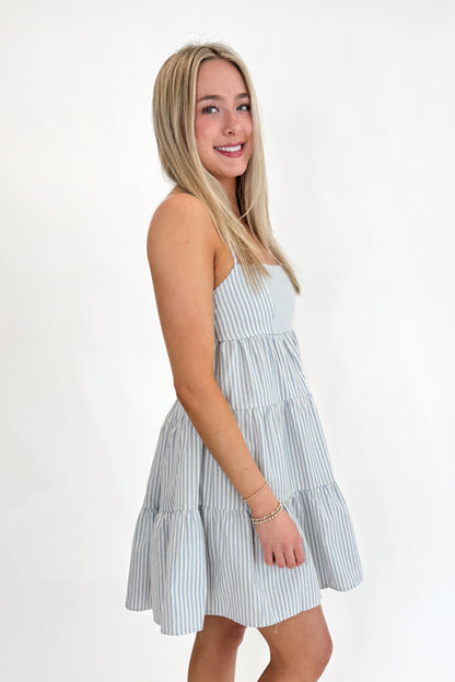 Side view of woman wearing Swagger Boutique’s Blue Striped Sleeveless Tiered Mini Dress with sandals against white studio background.