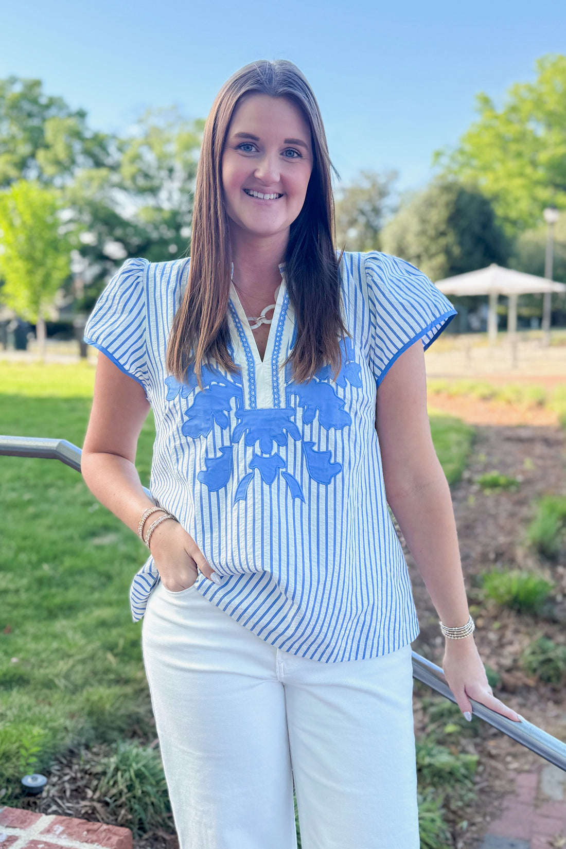 Front view of woman wearing Blue Striped &amp; Embroidered Flutter Sleeve Top with jeans outside at a park.