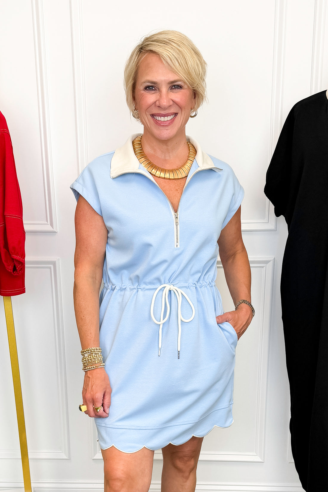 Front view of woman wearing Swagger Boutique’s Blue With White Lining Scallop Hem 1/4 Zip Dress with sneakers with clothing hanging in the background.