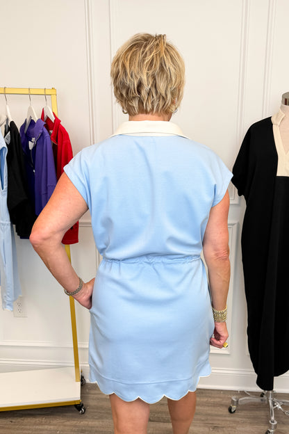 Back view of woman wearing Swagger Boutique’s Blue With White Lining Scallop Hem 1/4 Zip Dress with sneakers with clothing hanging in the background.