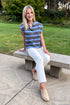 Front view of woman wearing Swagger Boutique’s Blue & Brown Striped Sleeveless Sweater by Entro with jeans sitting on a bench outside.