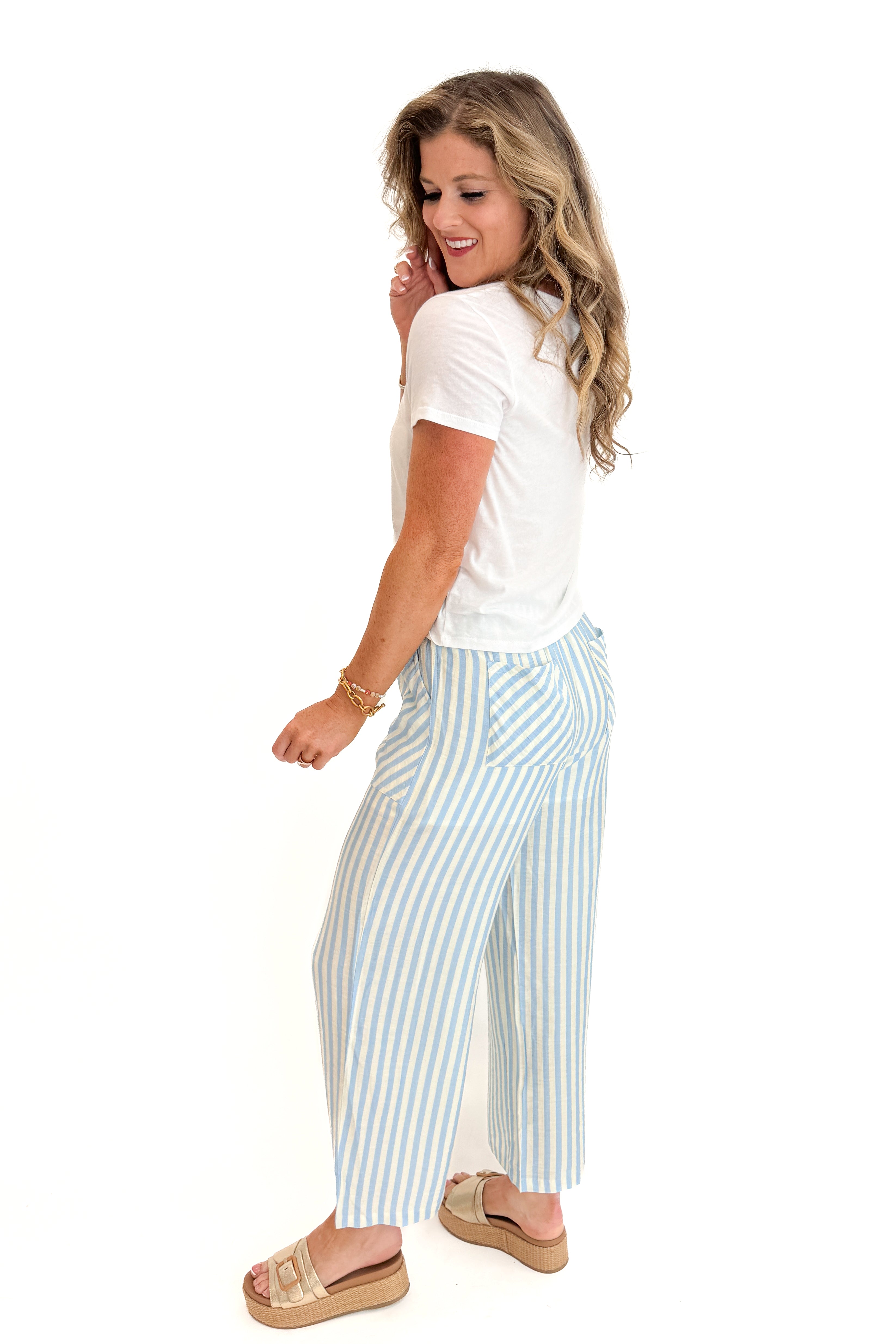 Side view of woman wearing Swagger Boutique’s Blue &amp; Cream Striped Pants with white top against white studio background.